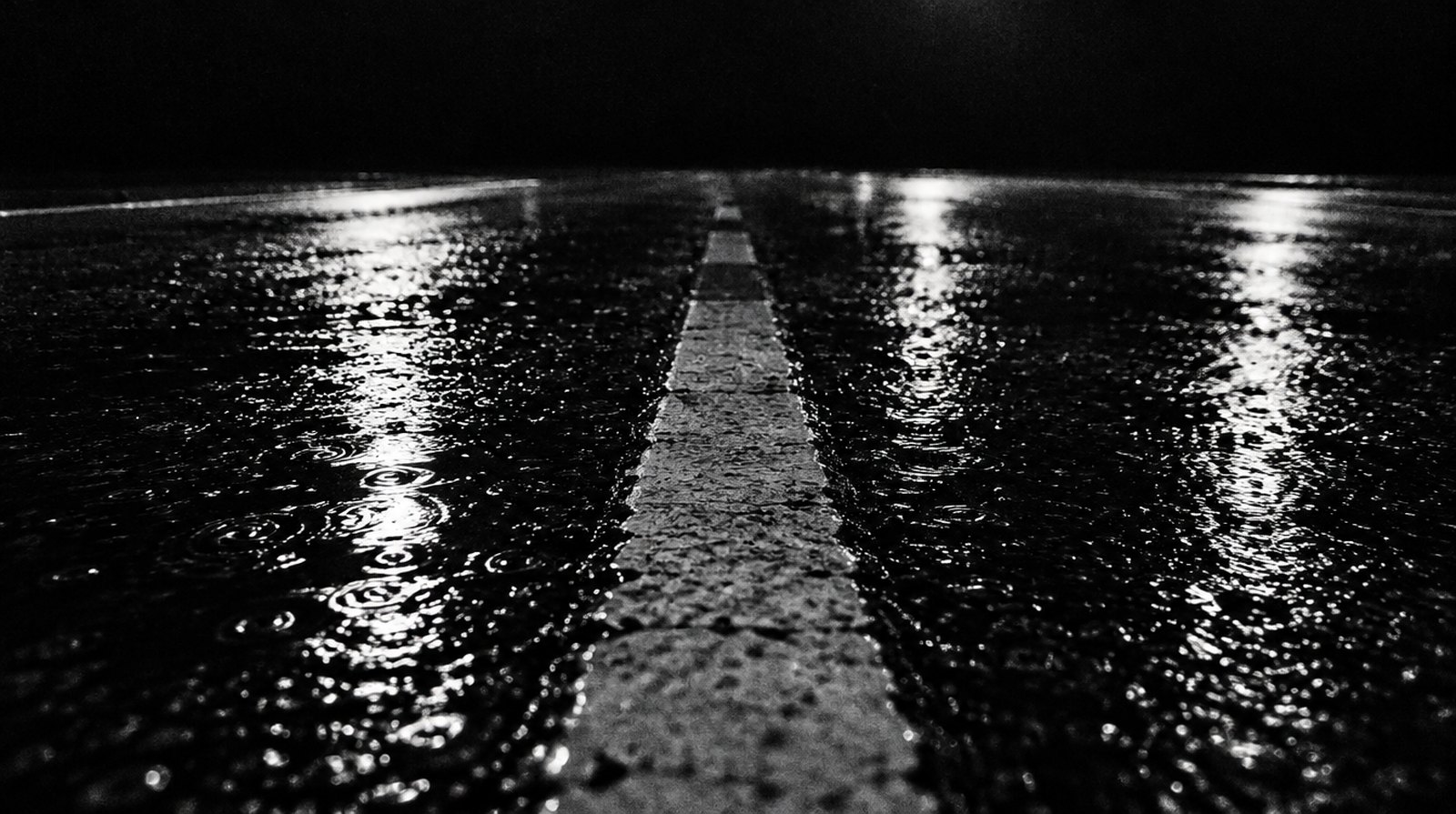 KI-generierte Darstellung: nasser Asphalt bei Nacht aus flachem Blickwinkel, eine Fahrbahnmarkierung läuft in die Tiefe, Lichtreflexe und Regenringe auf der Oberfläche